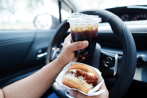 Driver holds soda in one hand and a sandwich in the other while trying to steer the wheel of a car, putting her at risk of a serious car accident.