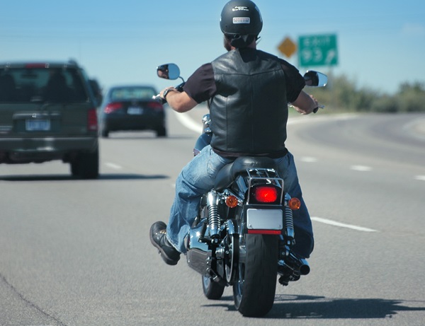 Motorcyclist wearing a helmet, leather vest, and jeans riding alone on a highway under a clear blue sky, with cars visible ahead in adjacent lanes.
