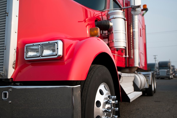 Close-up view of the front side of a red semi-truck parked on a road, with other trucks visible in the background.