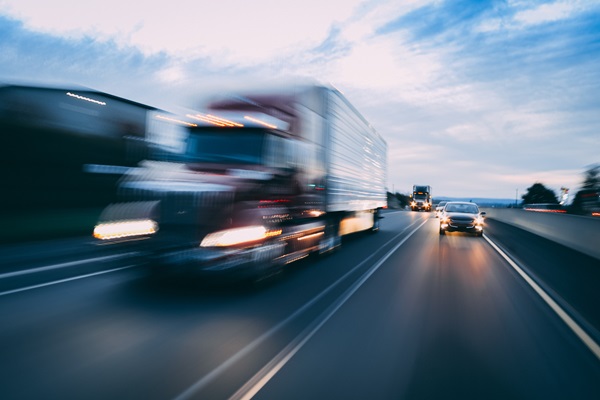 Blurred view of a semi-truck and several cars traveling at high speed on a multi-lane highway.